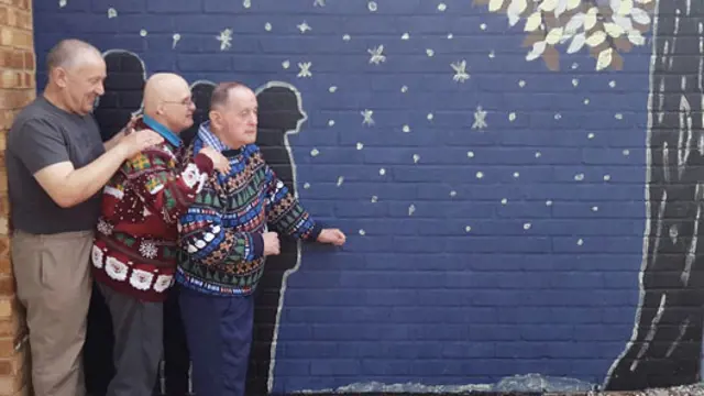 Three men standing in front of a painted wall decorated with stars and a tree